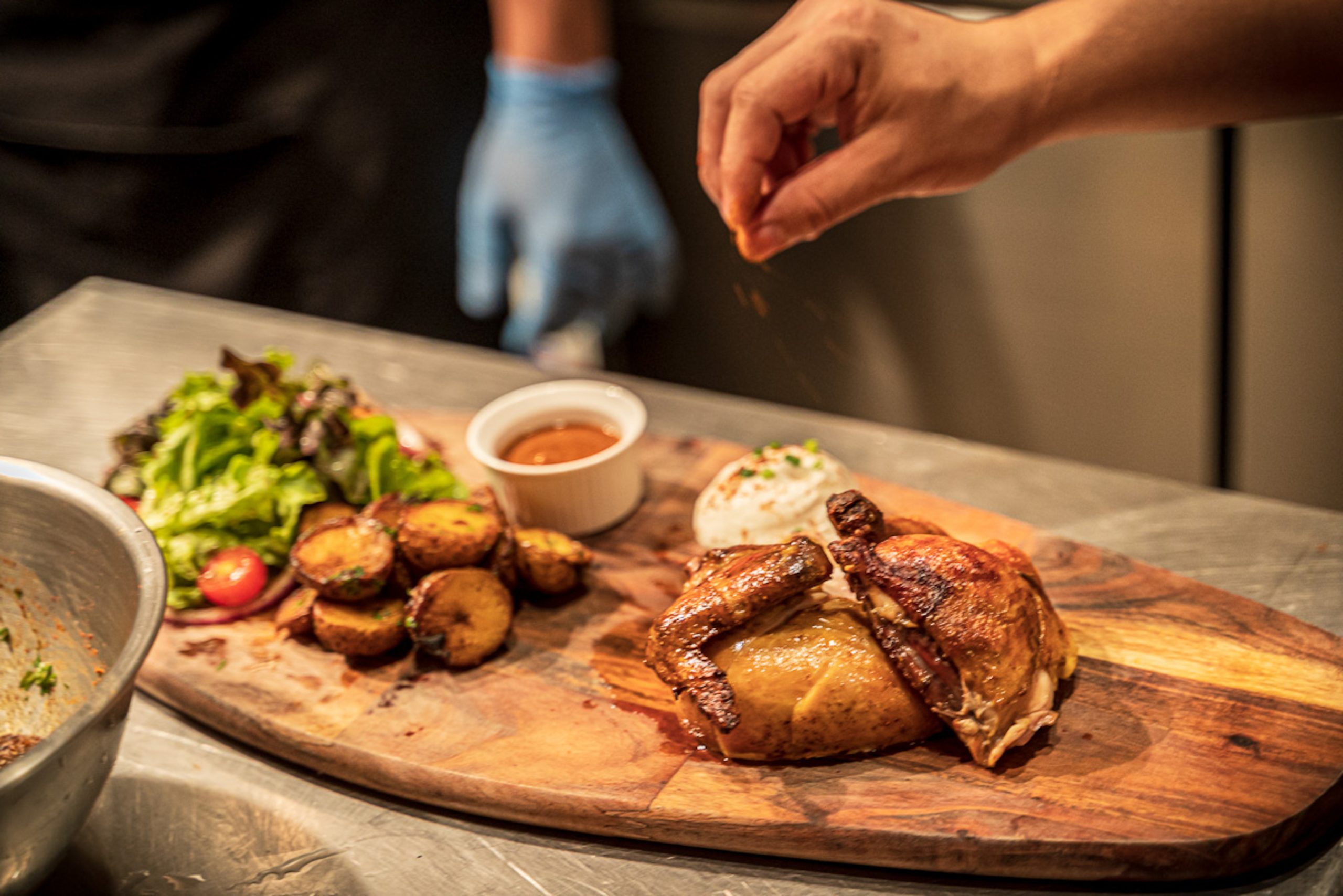 Commercial food photography of a chef seasoning plated rotisserie chicken with roasted potatoes, salad and sauce.