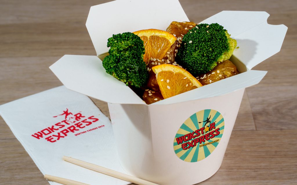 Orange-glazed chicken with broccoli, orange wedges and sesame in a takeaway box.